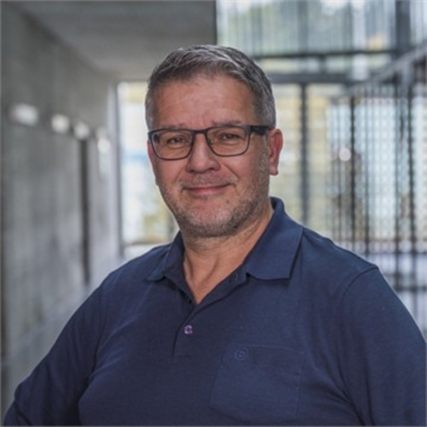A smiling man in glasses and a polo shirt stands in a modern, well-lit hallway.