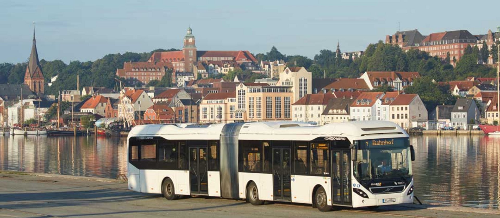 Modern articulated bus by waterfront with historic town buildings in background